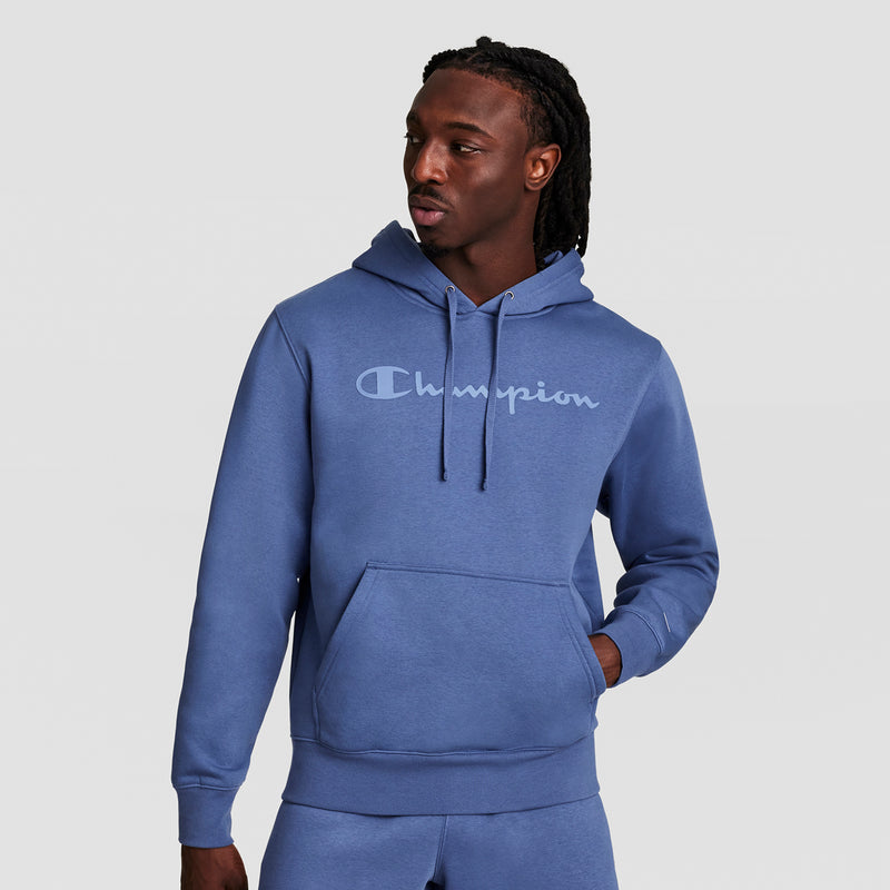 A man wears a blue Champion Powerblend Hoodie with a tonal script logo and matching fleece pants, standing against a plain light background, looking left with one hand in his pocket.