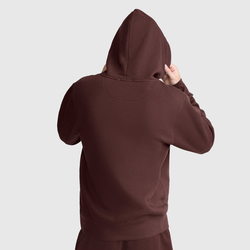 A person is shown from the back wearing a brown Champion Powerblend Hoodie with a tonal script logo, hood up and holding the sides of the hood. The Champion script appears on the sleeve, and the background is plain light gray.