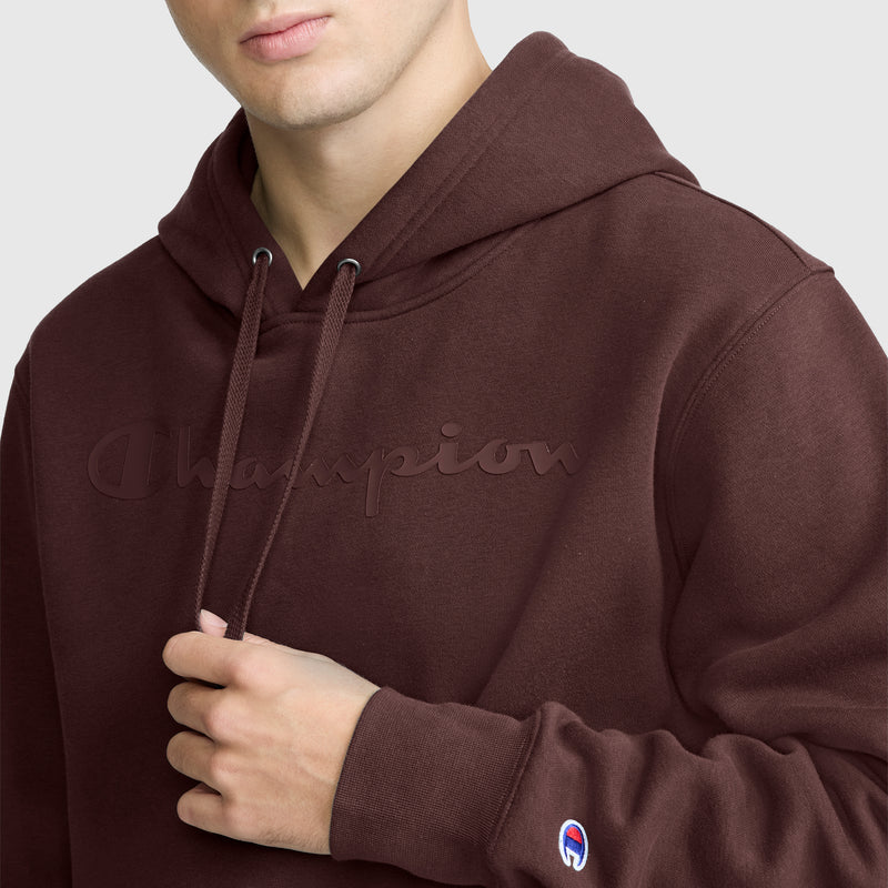 A person wearing a brown Champion Powerblend Hoodie with a Tonal Script Logo holds one of the hoodie’s drawstrings. The subtle Champion script is visible on the chest, while a small Champion patch on the sleeve adds a signature touch to this classic style.