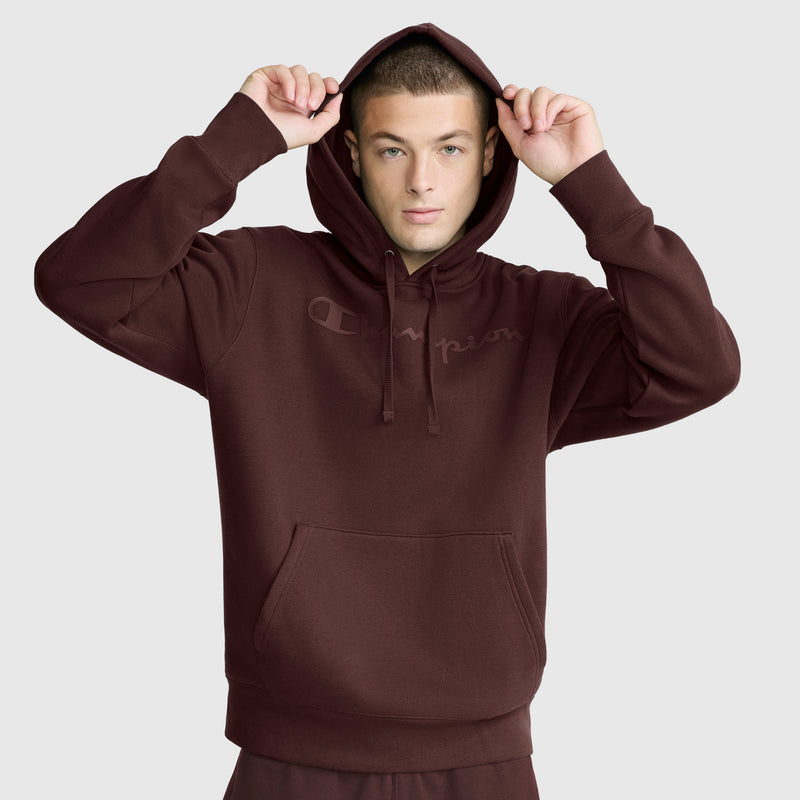 A person wearing a maroon Champion Powerblend Hoodie, Tonal Script Logo, with the hood up and holding the hood with both hands, stands against a plain light gray background.