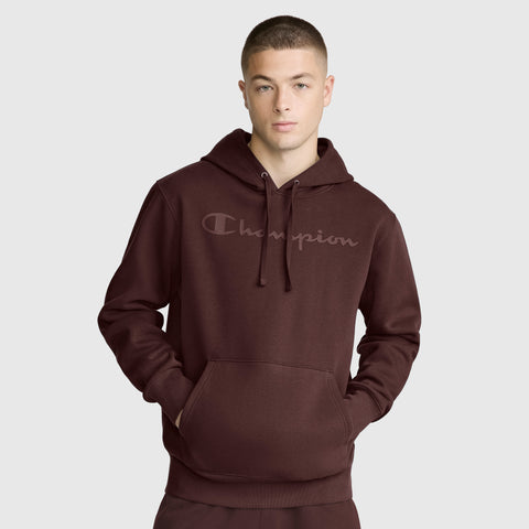 A person wearing the Champion Powerblend Hoodie with a tonal script logo, featuring a front pocket and drawstring hood, stands against a plain light gray background.