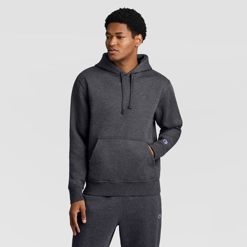 A man wearing a Champion Powerblend Hoodie, C Logo, in dark gray, along with matching sweatpants stands facing slightly to the left with one hand in his pocket against a plain light gray background.