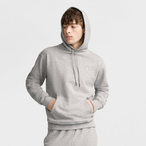 Young person in a light gray Champion Powerblend Hoodie, C Logo and matching sweatpants stands with hands in front pocket, looking to the side against a plain light background.