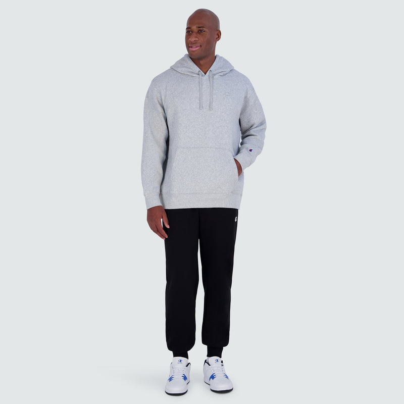 A man wearing a light gray Champion Powerblend Hoodie, C Logo - Big & Tall with a front pocket, black sweatpants, and white sneakers.