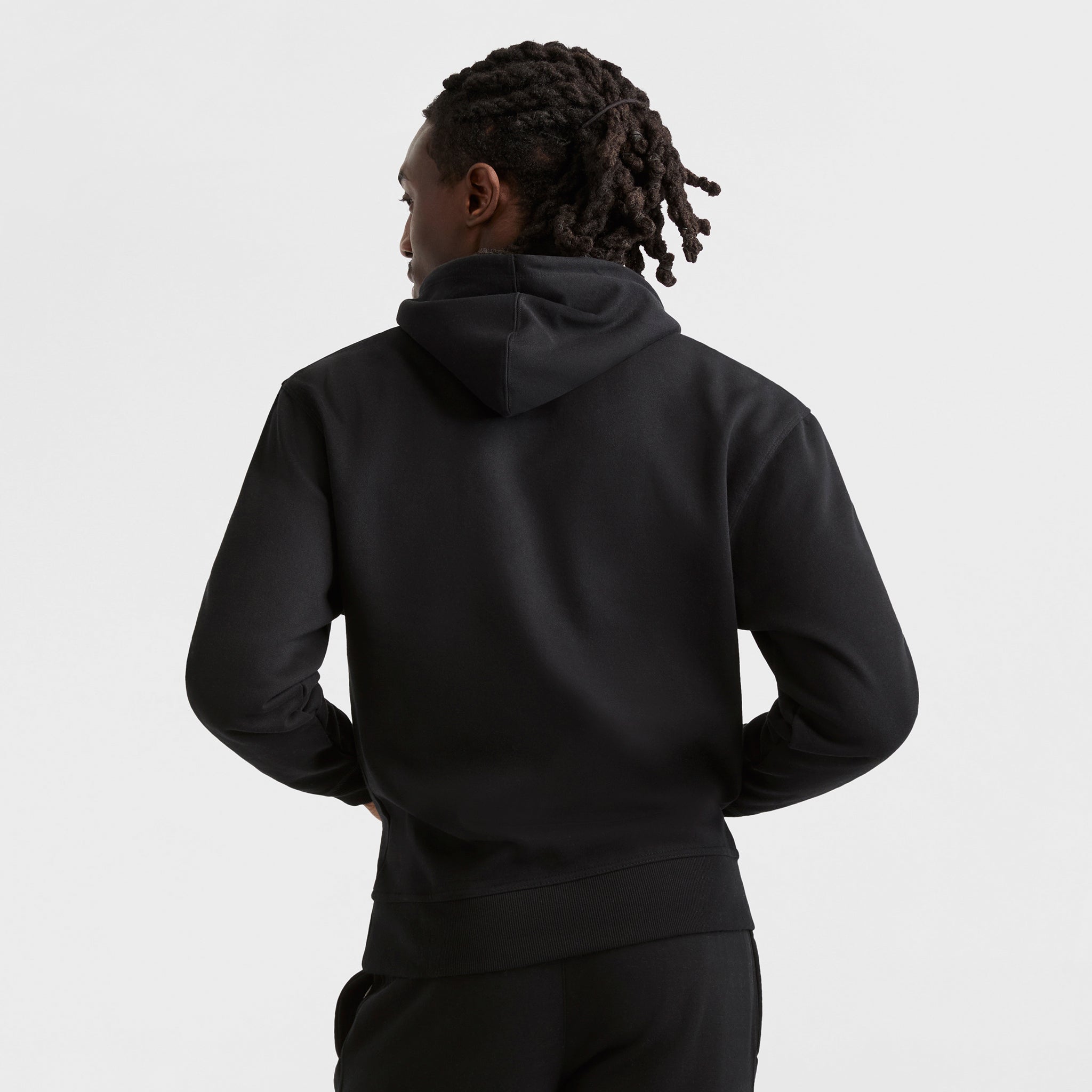 A person with medium-length dreadlocks faces away from the camera, wearing a plain black Champion Powerblend Hoodie, C Logo, and black pants against a white background.