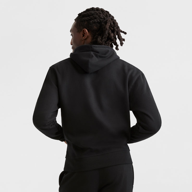 A person with medium-length dreadlocks faces away from the camera, wearing a plain black Champion Powerblend Hoodie, C Logo, and black pants against a white background.