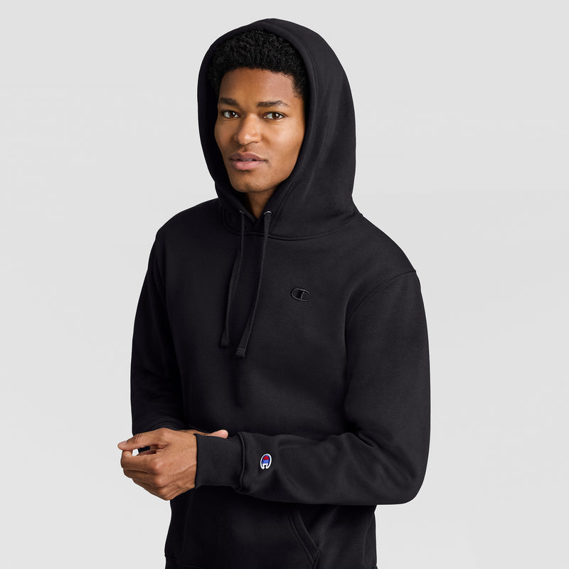 A person wearing a black Champion Powerblend Hoodie, C Logo, stands against a plain light gray background, looking at the camera with a slight smile and hands held together in front.