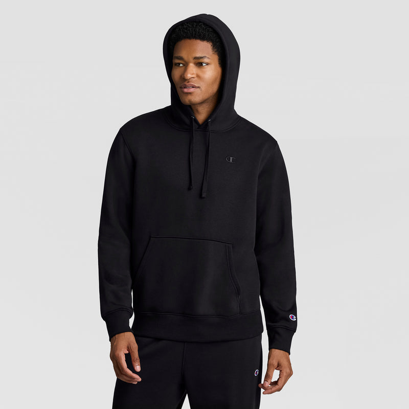 A person wearing a black Champion Powerblend Hoodie, C Logo, and matching black pants stands against a plain light gray background, looking slightly to the side with one hand in their pocket.