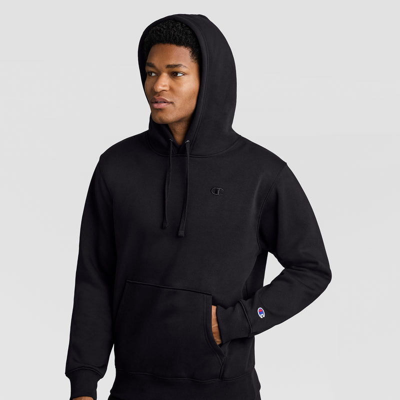 A person wearing a black Champion Powerblend Hoodie, C Logo, with the hood up stands against a plain light background, one hand casually tucked in the front pocket and a neutral expression.