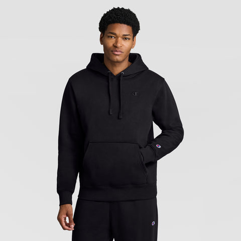 A person wearing a Champion Powerblend Hoodie, C Logo in black and black sweatpants stands against a plain light gray background, with one hand in the front pocket of the men’s hooded sweatshirt.