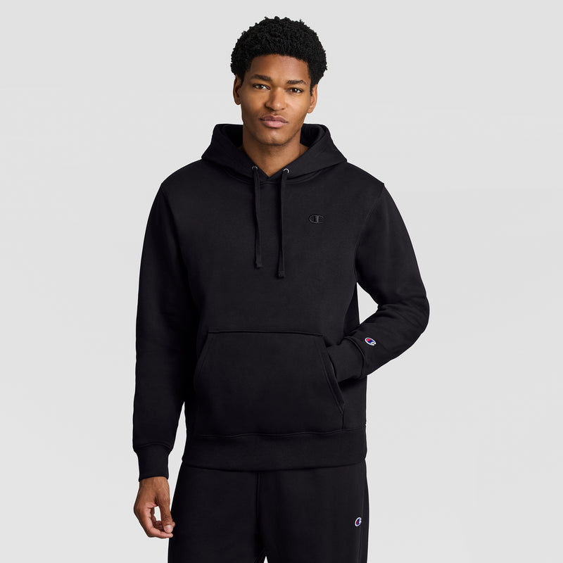 A person wearing a Champion Powerblend Hoodie, C Logo in black and black sweatpants stands against a plain light gray background, with one hand in the front pocket of the men’s hooded sweatshirt.
