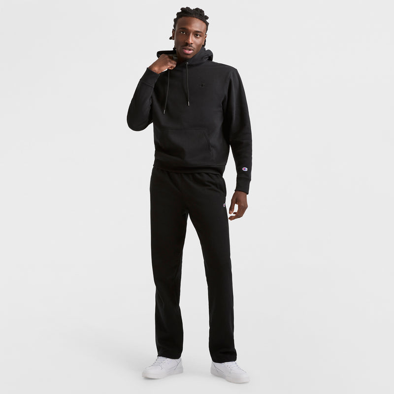 A man stands against a plain white background wearing the Champion Powerblend Hoodie, C Logo, with black pants and white sneakers. He holds one hoodie string in his right hand and looks at the camera with a neutral expression.