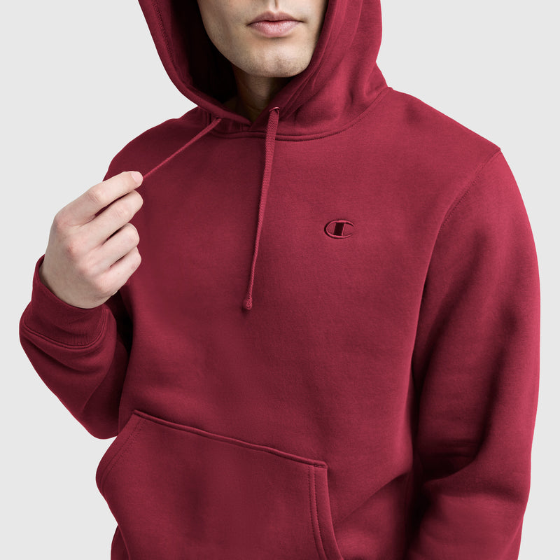 A person in a red Champion Powerblend Hoodie, C Logo with front pocket and drawstrings adjusts the hood. The C logo is visible on the left chest against a plain, light-colored background.