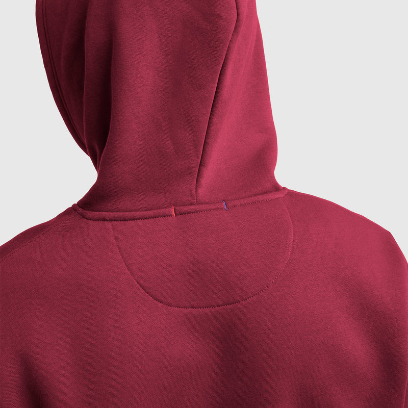 A close-up of the back of a maroon Powerblend Hoodie, C Logo by Champion shows detailed stitching and two small colored stitches—one red, one blue—on the seam below the hood.
