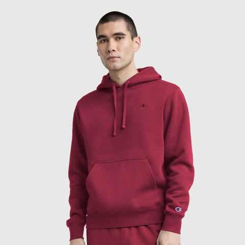 A person wears the Champion Powerblend Hoodie, C Logo in red with matching sweatpants, standing against a plain light gray background and looking at the camera neutrally.