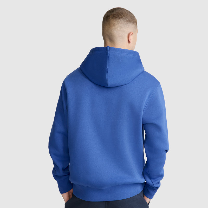 A person stands facing away from the camera against a plain light background, wearing a bright blue Champion Powerblend Hoodie, C Logo, with the hood down.