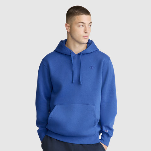 A young man wearing the Champion Powerblend Hoodie with C Logo, featuring a front pocket and drawstrings, stands against a plain light gray background, looking to his left with his hands in his pockets.
