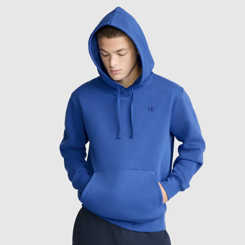 A person wearing the Champion Powerblend Hoodie, C Logo in blue stands against a plain light gray background with hands in the front pocket, looking down.