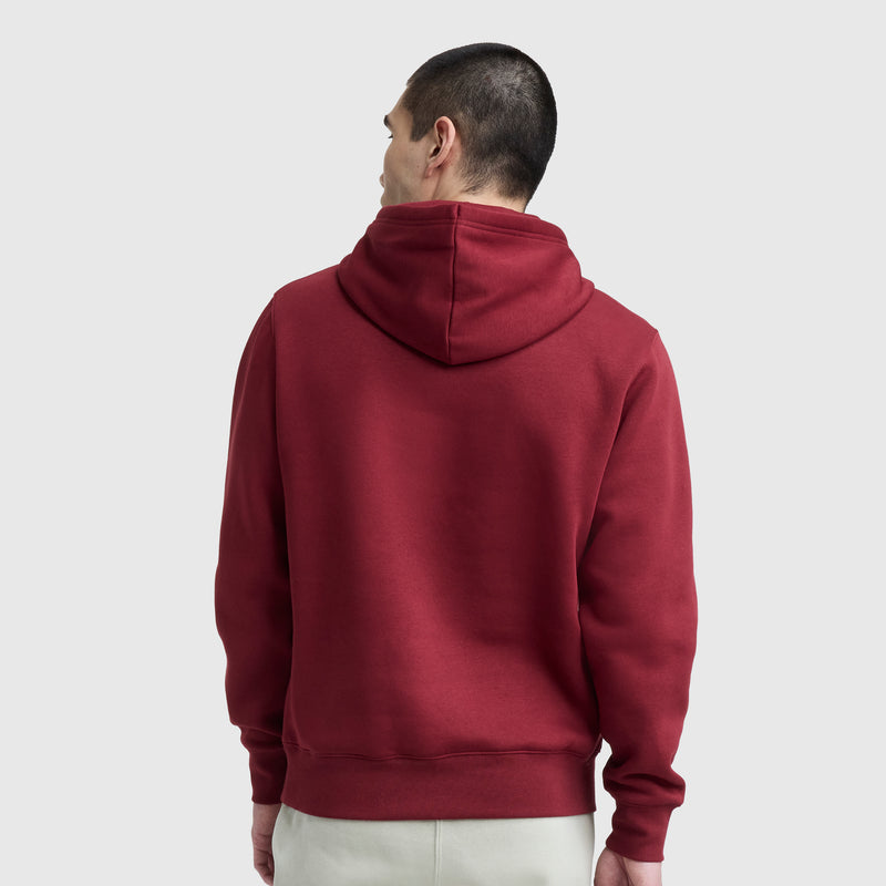 A person with short dark hair, seen from behind, wears a maroon Champion Powerblend Hoodie, C Logo, and light-colored pants while standing against a plain light gray background.