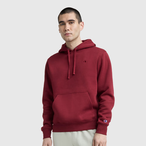 A person is wearing the Champion Powerblend Hoodie, C Logo in maroon with a front pocket and drawstrings, paired with light-colored pants, against a plain light gray background.