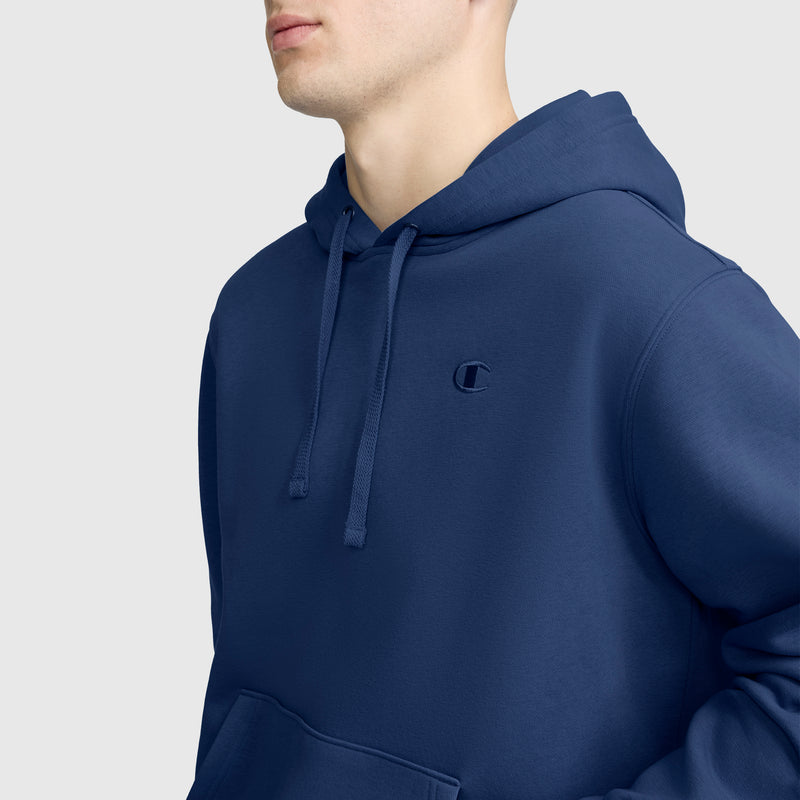 A person wearing a blue Powerblend Hoodie, C Logo by Champion stands against a plain light gray background. The photo is cropped at the nose and above the waist, highlighting the fleece hoodie details.
