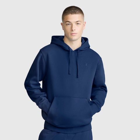 A person wearing a plain navy blue Champion Powerblend Hoodie, C Logo and matching sweatpants stands with hands in the hoodie pocket against a light gray background.