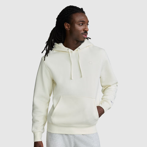 A man with long dark dreadlocks wears the Champion Powerblend Hoodie, C Logo, in a cream color, paired with light gray sweatpants. He stands against a plain gray background, looking to his right with one hand in his pocket.