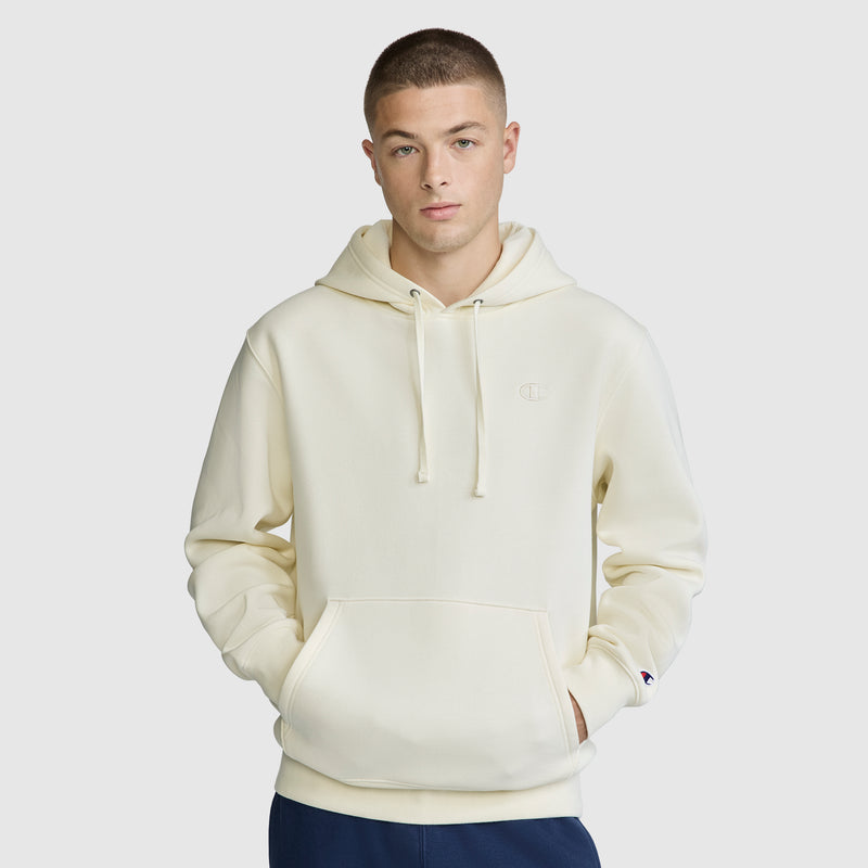 A person wearing a cream-colored Champion Powerblend Hoodie, C Logo, featuring a front pocket and drawstrings, stands facing forward with hands in the pocket against a plain light gray background.