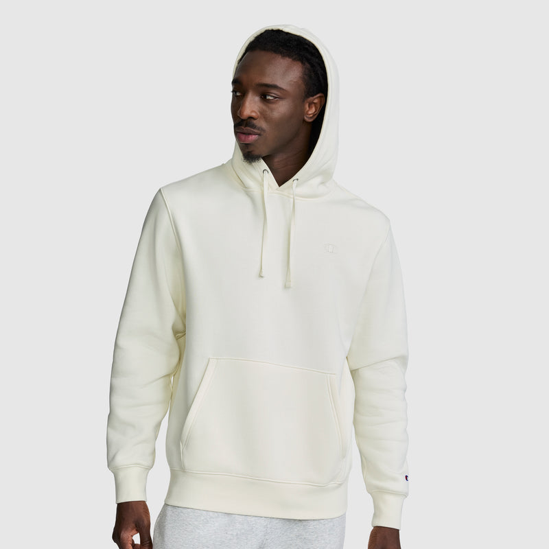 A man wearing a cream-colored Champion Powerblend Hoodie, C Logo, featuring a front pocket and hood up, paired with light gray sweatpants, stands against a plain gray background.
