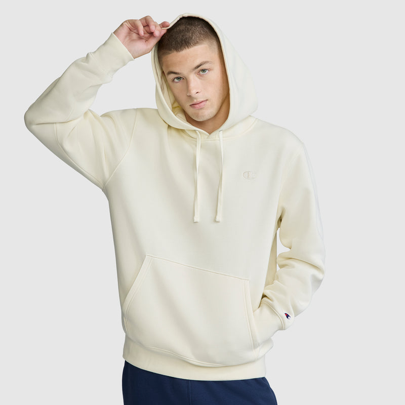 A man wearing a light cream Champion Powerblend Hoodie with the hood up holds the edge of the hood with one hand. He has short hair and is looking at the camera with a neutral expression, against a plain light background. The Champion Powerblend Hoodie, featuring the signature C Logo, delivers both comfort and style for any casual occasion.