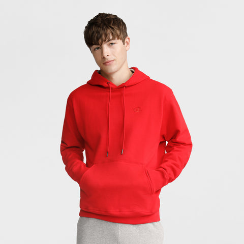 A young man with short brown hair wears a Champion Powerblend Hoodie, C Logo, in bright red with a front pocket and drawstrings, paired with light gray pants. He stands with his hands in the pocket against a plain light background.
