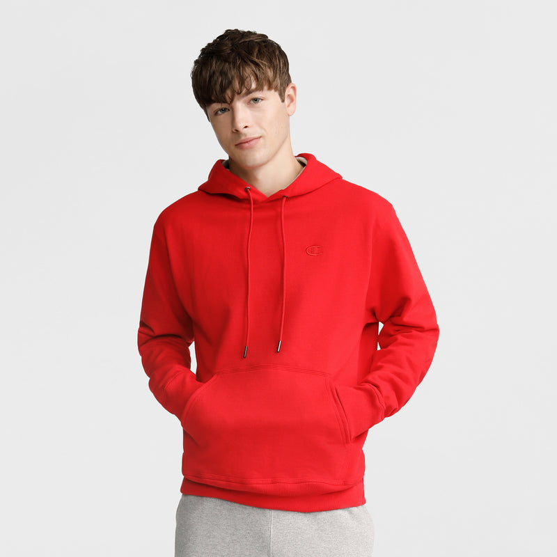 A young man with short brown hair wears a Champion Powerblend Hoodie, C Logo, in bright red with a front pocket and drawstrings, paired with light gray pants. He stands with his hands in the pocket against a plain light background.