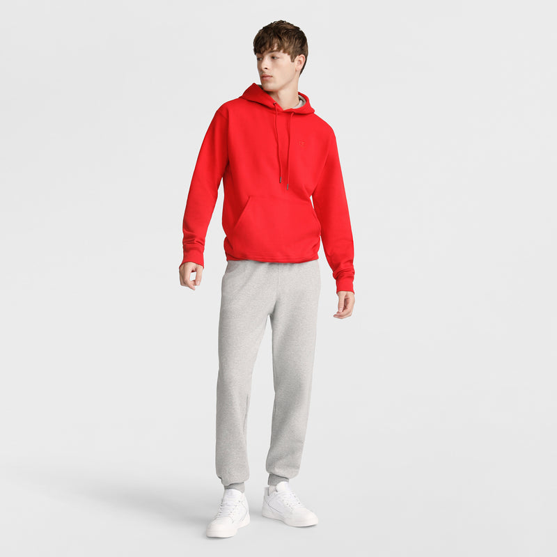 A person stands against a plain background wearing a Champion Powerblend Hoodie, C Logo in bright red, paired with light gray sweatpants and white sneakers, looking slightly to the side with a relaxed posture.