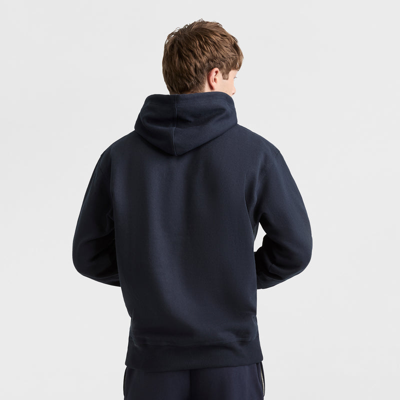 A person with light brown hair stands with their back to the camera, wearing a dark navy Champion Powerblend Hoodie, C Logo, and matching pants against a plain white background.