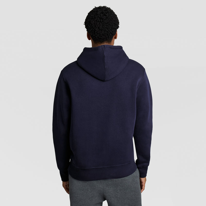 A person stands facing away against a light gray background, wearing gray pants and a dark blue Champion Powerblend Hoodie with the C Logo.