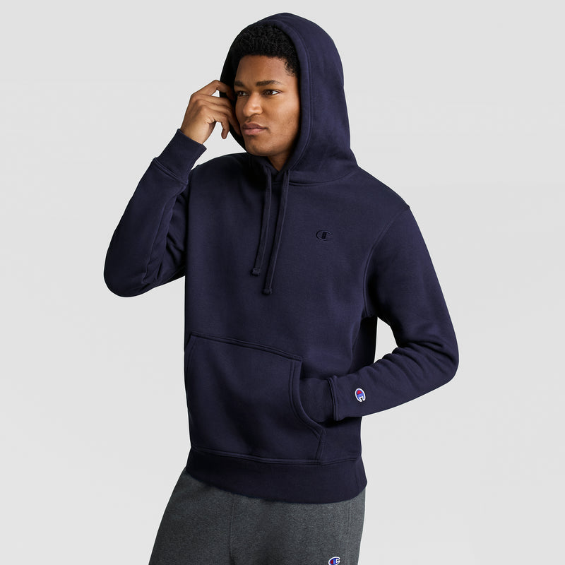 A man wears a dark navy blue Champion Powerblend Hoodie, C Logo, with the hood pulled up, posing with one hand near his face and the other in the kangaroo pocket. The men’s sweatshirt features a small tonal “C” logo on the chest and a colorful Champion patch on the sleeve.