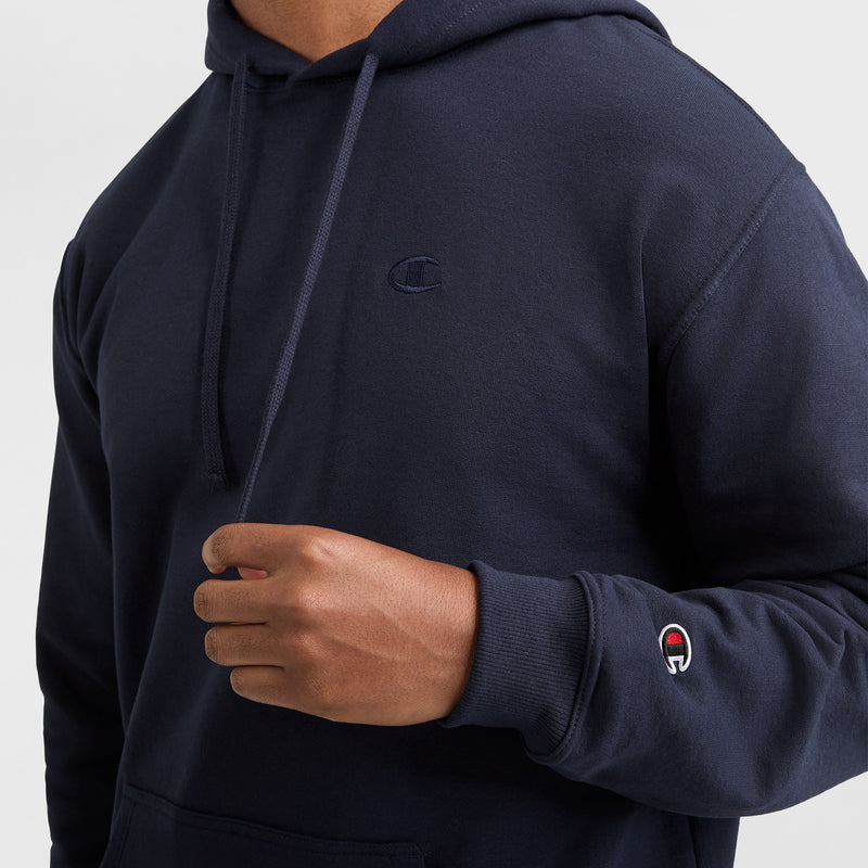 A person wearing a navy blue Champion Powerblend Hoodie, C Logo, with one hand raised near the chest. The cozy hoodie features visible Champion branding on the chest and sleeve.