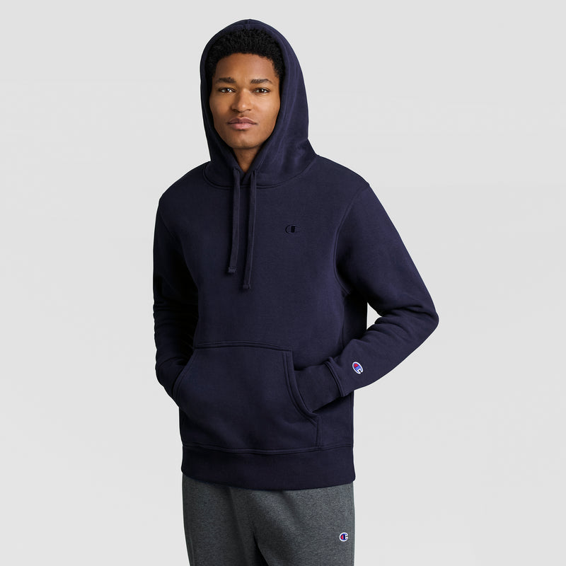 A person wearing the Champion Powerblend Hoodie, C Logo in dark blue and gray sweatpants stands with hands in the hoodie pocket, facing forward against a plain light background.