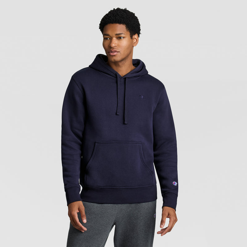A man wearing the Champion Powerblend Hoodie, C Logo in navy blue stands against a plain light gray background. He has short curly hair, is dressed in dark gray sweatpants, and looks slightly to the side with a neutral expression.