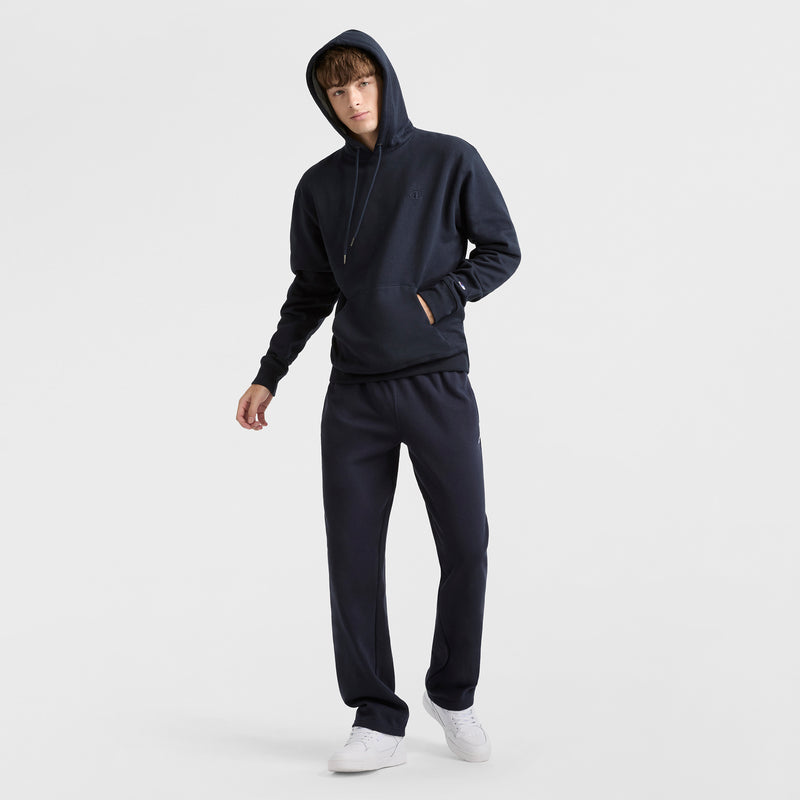 A person in a black Champion Powerblend Hoodie, C Logo, and matching black sweatpants stands against a white background, one hand in their pocket and the other arm relaxed, paired with white sneakers.