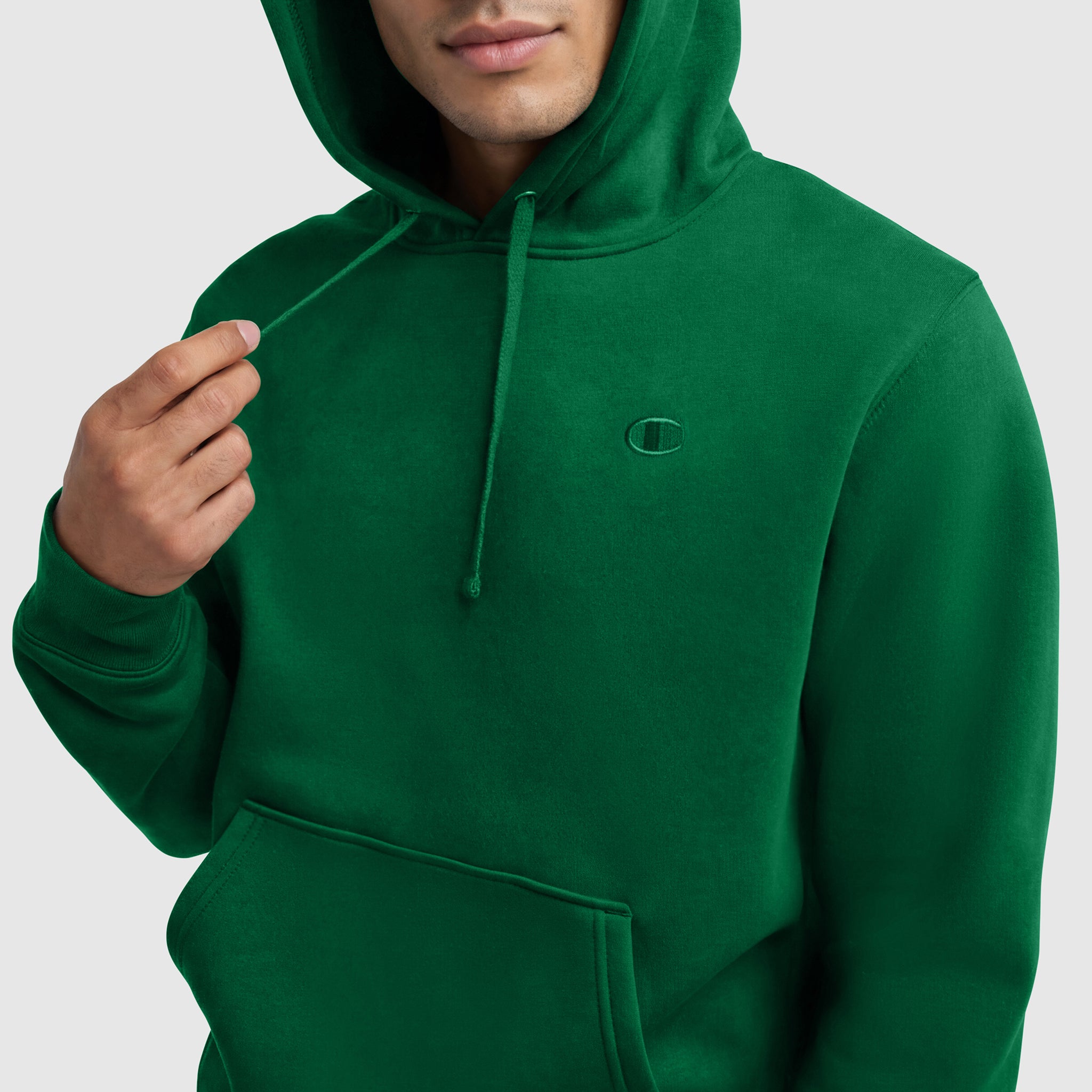 A person wearing the Champion Powerblend Hoodie, C Logo in green with a front pocket holds a drawstring. The image is cropped at the upper chest, showing only the lower half of their face and torso.