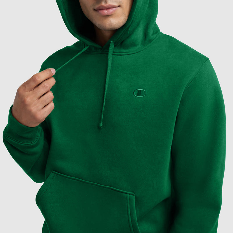A person wearing the Champion Powerblend Hoodie, C Logo in green with a front pocket holds a drawstring. The image is cropped at the upper chest, showing only the lower half of their face and torso.