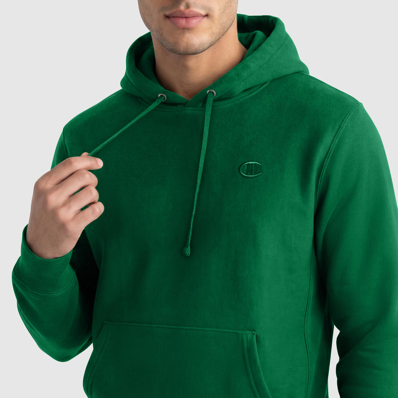 A person wears a bright green Champion Powerblend Hoodie, featuring a small embroidered C logo on the chest, and holds one drawstring in their left hand. The men's sweatshirt partially reveals their face against a plain light background.