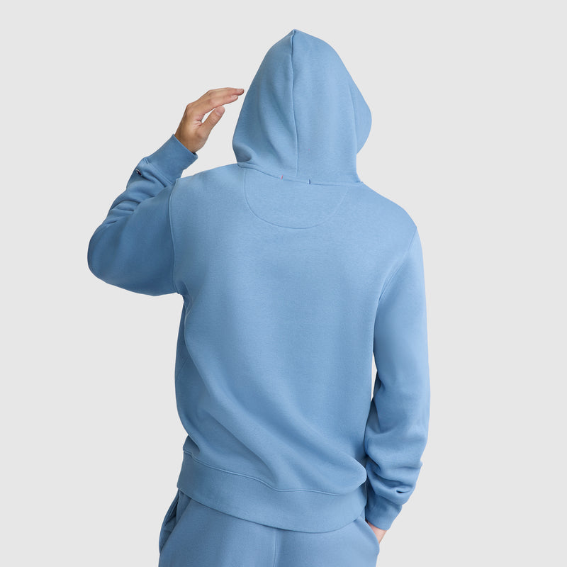 A person wearing the Champion Powerblend Hoodie, C Logo in light blue, along with matching pants, stands with their back to the camera and raises one hand to adjust the hood. The background is plain white.