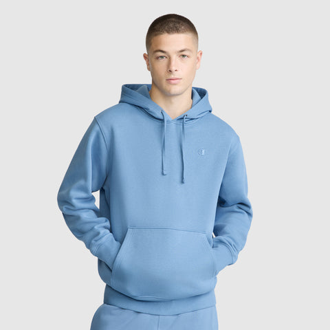 A person wearing a light blue Champion Powerblend Hoodie, C Logo, with their hands in the front pocket, standing against a plain light gray background.