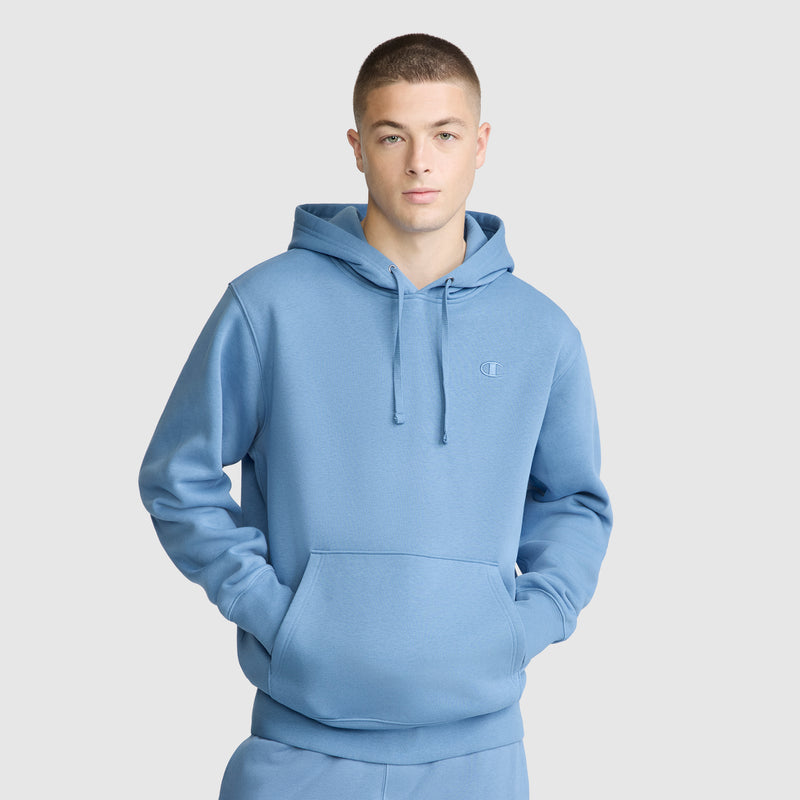 A person wearing a light blue Champion Powerblend Hoodie, C Logo, with their hands in the front pocket, standing against a plain light gray background.