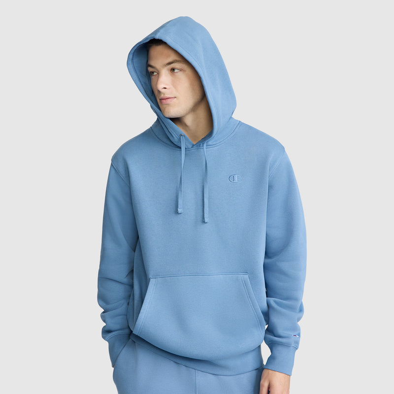A person wearing the Champion Powerblend Hoodie, C Logo in light blue with the hood up and matching sweatpants, standing against a plain light background and looking to the side with one hand in the pocket.