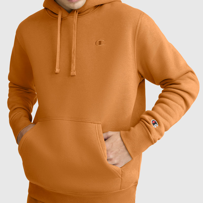 A person wearing a Champion Powerblend Hoodie, C Logo in orange with their hands in the front pocket, displaying Champion branding on the chest and left sleeve, set against a plain light background.