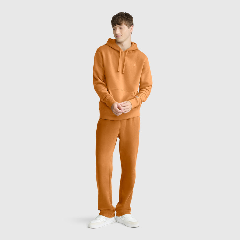 A person stands wearing a Champion Powerblend Hoodie, C Logo in orange with matching sweatpants and white sneakers, posed against a plain light gray background.