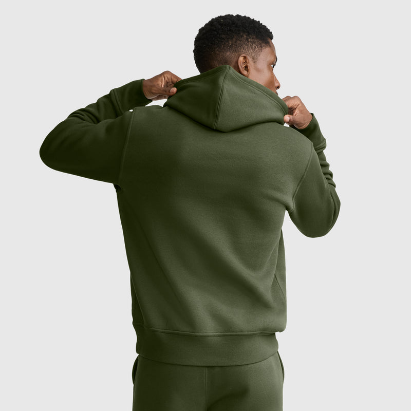 A person wearing a Champion Powerblend Hoodie, C Logo in olive green with matching pants is seen from behind, holding up the hooded sweatshirt with both hands against a plain, light gray background.